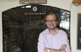 Ein Mann mit Brille und rosa Hemd steht lächelnd vor einem Schild mit der Aufschrift 'Martinshof'.