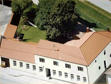 Luftaufnahme eines Gebäudekomplexes mit rotem Dach und Innenhof mit Bäumen und Spielplatz.