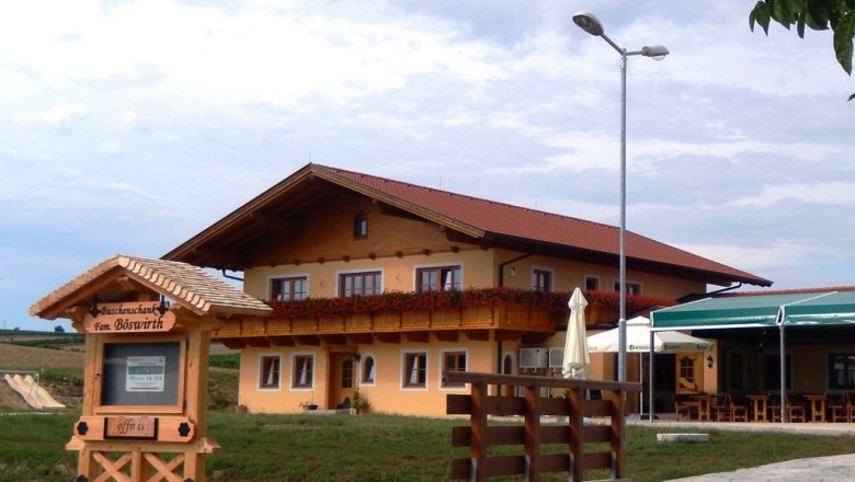 Ein traditionelles Gasthaus mit Holzbalkon und Blumendekoration, daneben ein Schild mit der Aufschrift 'Buschenschank Fam. Böswirth'.