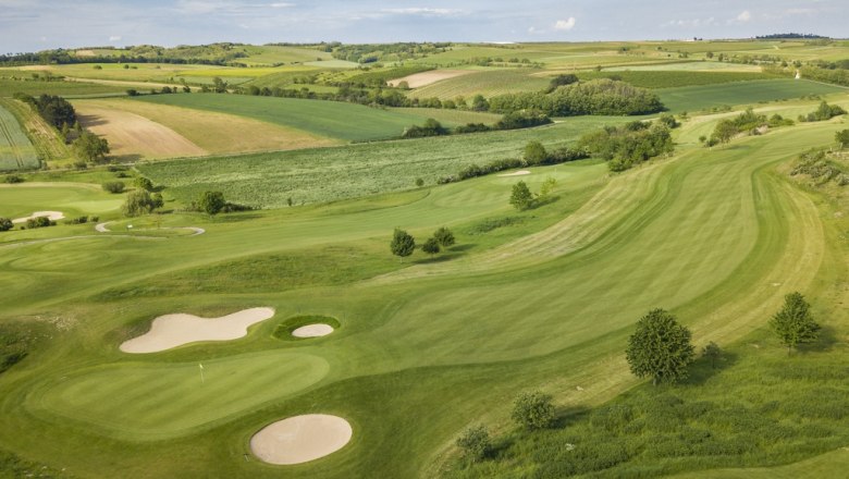 Luftaufnahme eines Golfplatzes mit grünen Fairways und Sandbunkern, umgeben von Feldern und Hügeln.