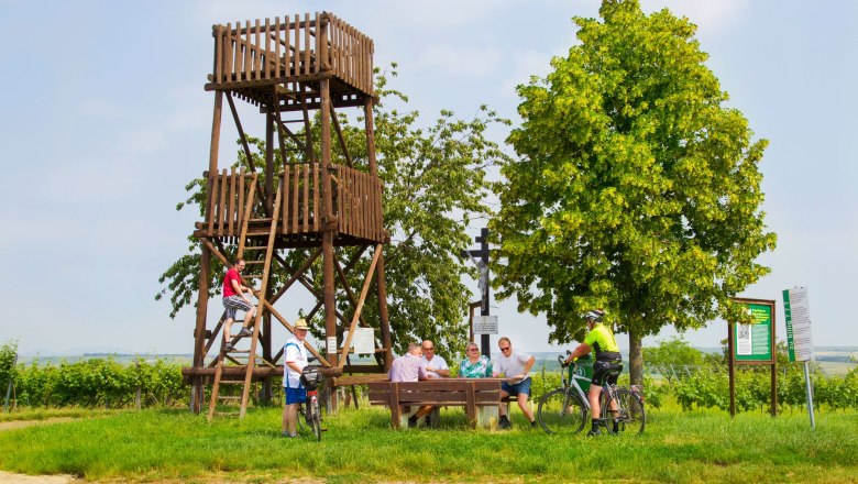 Menschen entspannen an einem Holzturm und Tisch im Freien, umgeben von B&auml;umen und Schildern.