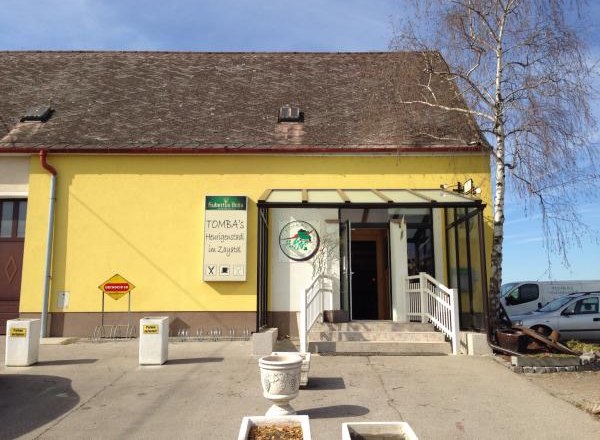 Gelbes Gebäude mit Schild 'Tomba's Heurigenschenke'. Eingang mit Treppe und Glasüberdachung.