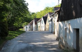 Reihe von Weinkellern in einer ländlichen Straße mit Bäumen im Hintergrund.