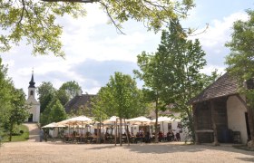 Ein Dorfwirtshaus im Museumsdorf mit Sonnenschirmen und einer Kirche im Hintergrund.