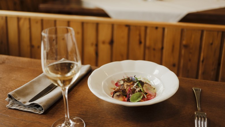 Ein Glas Wei&szlig;wein neben einem Teller mit einem dekorativen Gericht auf einem Holztisch.