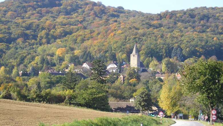 Eine malerische Landschaft mit einer Stra&szlig;e, die zu einem Dorf mit einer Kirche f&uuml;hrt, umgeben von bewaldeten H&uuml;geln im Herbst.