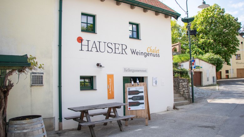 Weingenuss.Chalet, © Hintersteiner Gerald Lifedesign Außenansicht eines Gebäudes mit der Aufschrift 'Hauser Chalet Weingenuss'. Vor dem Gebäude steht ein Holztisch und ein Weinfass.