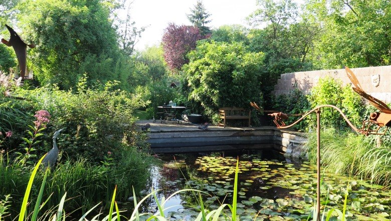 Ein idyllischer Garten mit Teich, Seerosen und üppiger Vegetation. Im Hintergrund stehen ein Tisch und Stühle auf einer Terrasse.