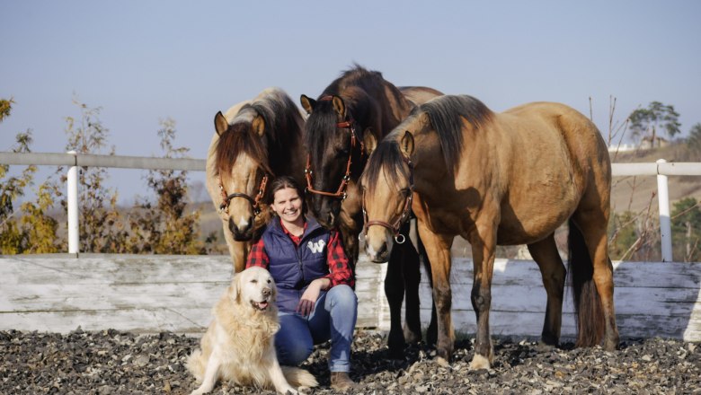Eine Frau sitzt l&auml;chelnd auf einem Reitplatz, umgeben von drei Pferden und einem Hund.