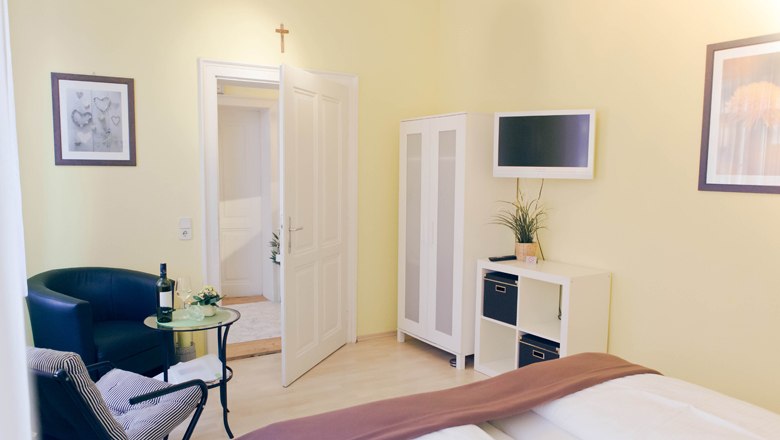 Helles Gästezimmer mit Bett, Sessel, Tisch, Schrank und Fernseher.