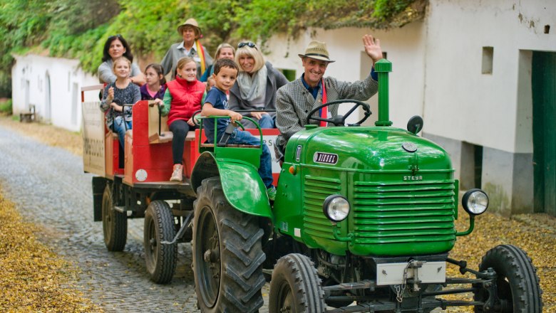 Grüner Traktor mit Anhänger voller Menschen auf einer gepflasterten Straße.