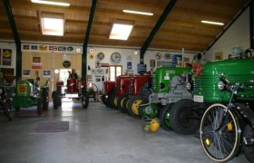 Innenansicht einer Scheune mit mehreren Oldtimer-Traktoren in verschiedenen Farben, umgeben von Retro-Werbeschildern an den W&auml;nden.