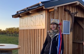 Eine Person mit Hut und Flasche steht vor einer Holzh&uuml;tte mit der Aufschrift 'Hauser'.