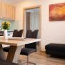Moderne K&uuml;che mit Holzm&ouml;beln, Esstisch, St&uuml;hlen und einem Sofa mit orangefarbenen Kissen.