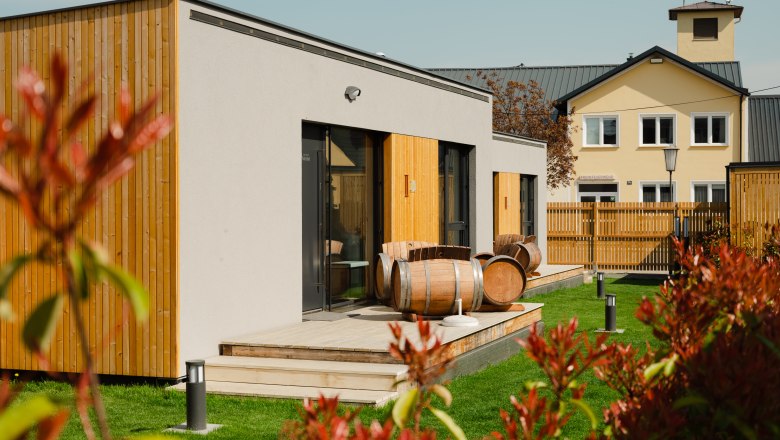 Moderne Appartements mit Holzverkleidung und Weinf&auml;ssern auf der Terrasse, umgeben von gr&uuml;nem Rasen und einem Zaun.