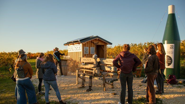 Gruppe von Menschen steht vor einem kleinen Holzh&auml;uschen in einem Weinberg bei Sonnenuntergang.