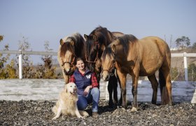 Eine Frau sitzt l&auml;chelnd auf einem Reitplatz, umgeben von drei Pferden und einem Hund.