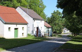 Eine ruhige Stra&szlig;e mit traditionellen H&auml;usern und gr&uuml;nen B&auml;umen an einem sonnigen Tag.