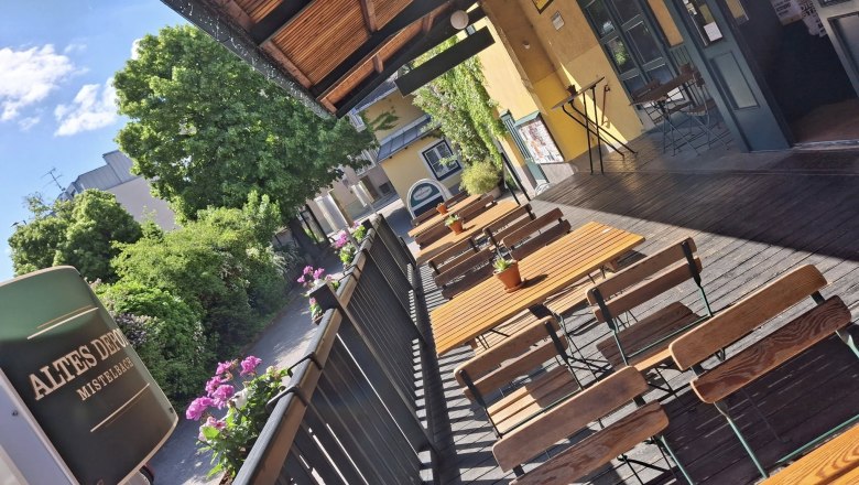 Eine sonnige Terrasse mit Holztischen und -stühlen, umgeben von Pflanzen und einem Gebäude mit gelber Fassade.