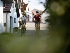 Zwei Radfahrer genie&szlig;en die sanfte Brise und die malerische Landschaft, w&auml;hrend sie durch die charmanten Gassen radeln. Umgeben von idyllischen Weinbergen und historischen Kellergassen, sp&uuml;ren sie die entspannte Atmosph&auml;re des Weinviertels.