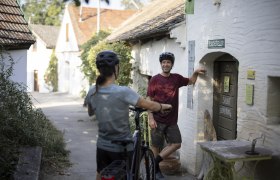 Zwei Radfahrer in einer Kellergasse, einer lehnt an einer Tür mit der Aufschrift 'Besucherkeller'.