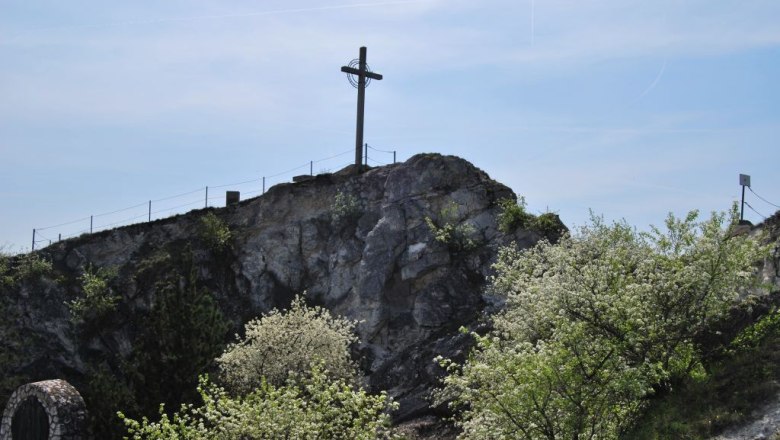 Ein Kreuz steht auf einem H&uuml;gel mit bl&uuml;henden B&auml;umen im Vordergrund.