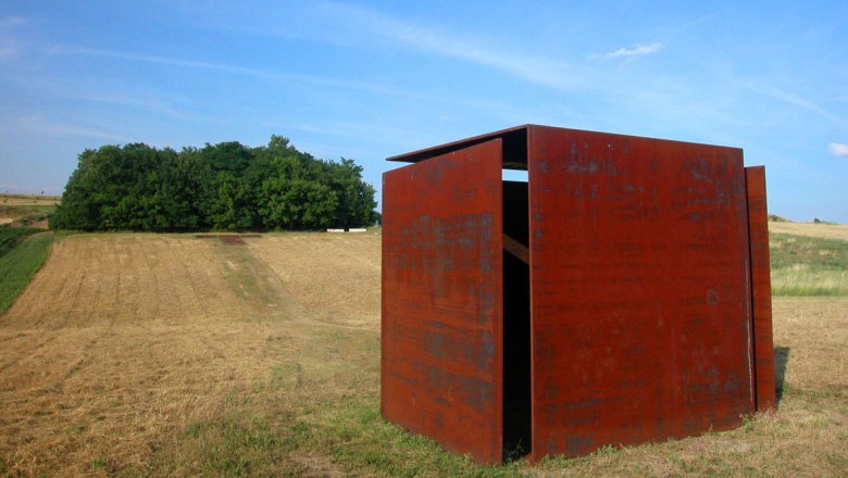 Eine rostige Metallskulptur steht auf einem Feld vor einem Wald unter blauem Himmel.