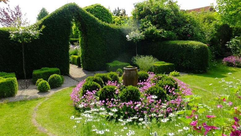 Ein gepflegter Garten mit Hecken, Blumenbeeten und einem großen Keramiktopf in der Mitte.