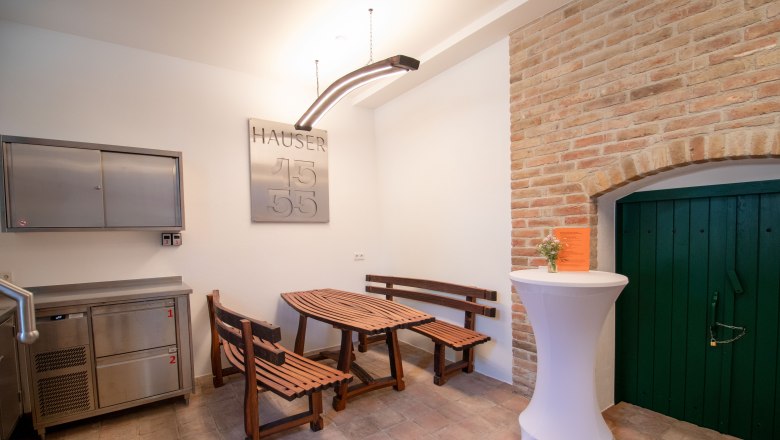 Ein rustikaler Raum mit Holzb&auml;nken, einem Holztisch, einem Stehtisch mit Blumen und einer gr&uuml;nen T&uuml;r in einer Ziegelwand.