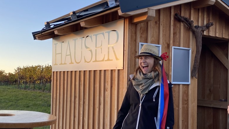 Eine Person mit Hut und Flasche steht vor einer Holzh&uuml;tte mit der Aufschrift 'Hauser'.