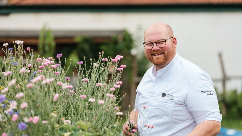 Ein lächelnder Mann in Kochkleidung steht neben blühenden Blumen im Freien.