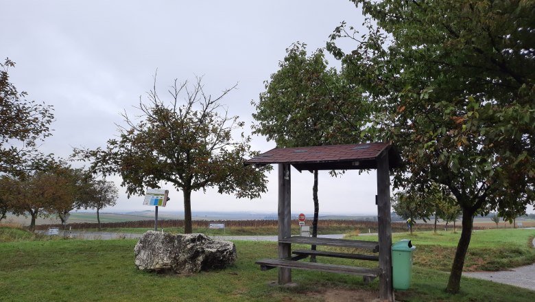 Ein Rastplatz mit Holzb&auml;nken und einem Tisch, umgeben von B&auml;umen und einer Wiese, unter einem bew&ouml;lkten Himmel.