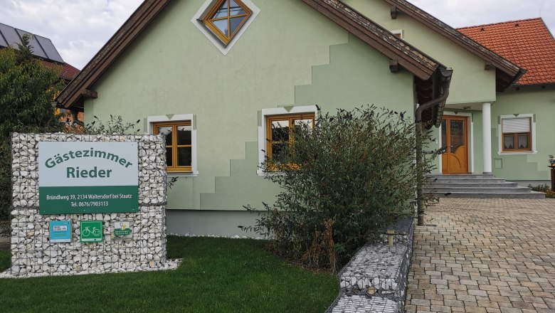 Gr&uuml;nes Haus mit Schild 'G&auml;stezimmer Rieder' im Vorgarten.