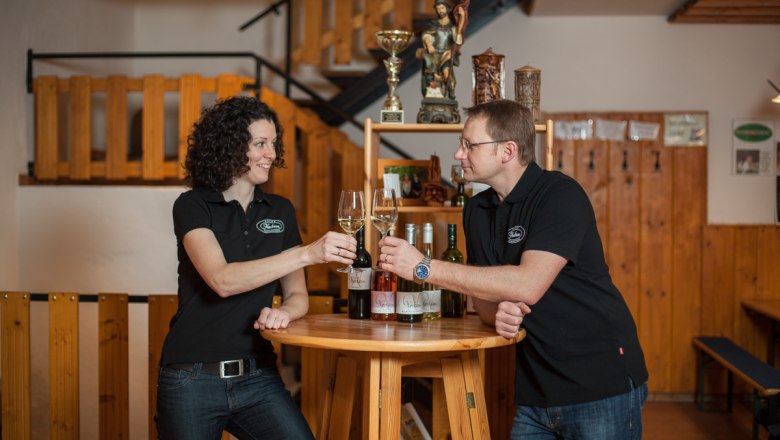 Zwei Personen stoßen mit Weingläsern an einem Holztisch an.
