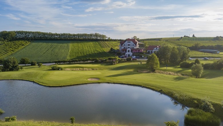 Landschaftsaufnahme eines Golfplatzes mit Teich und Clubhaus im Hintergrund.