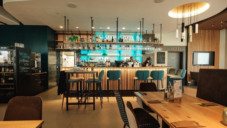 Moderner Barbereich mit Holztischen und blauen Barhockern, beleuchtet durch hängende Lampen.