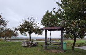 Ein Rastplatz mit Holzb&auml;nken und einem Tisch, umgeben von B&auml;umen und einer Wiese, unter einem bew&ouml;lkten Himmel.
